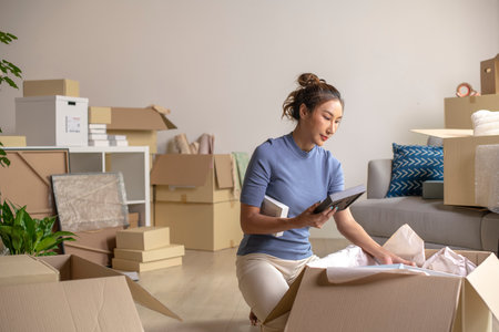 Young Asian woman moving into new apartment holding and unpacking her belongings with cardboard boxes.の写真素材