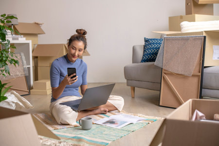 Young Asian woman sitting in new house using smartphone and laptop buying decorations online after moving in.の写真素材