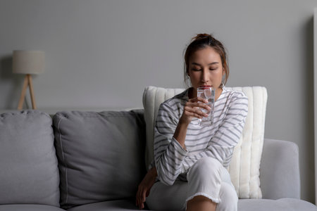 Asian woman drinking cold water while sitting on the sofa at home. Female drinking clean water...