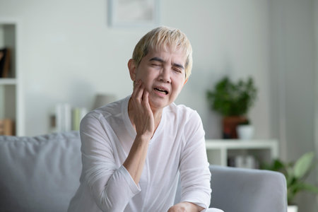 Asian mature woman with toothache. Elderly senior woman has toothache. Unhappy woman face in tooth pain sitting on sofa at home, feel sick unwell. Sad aged woman hand holding her chin. Adult suffering toothacheの写真素材