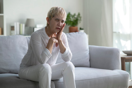 Senior Asian woman supports her cheek in pain from toothache while sitting on sofa at home.の写真素材