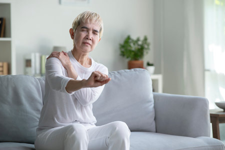 Elderly Asian woman at home with shoulder pain, Senior lady feeling muscles pain attack sitting on sofa in living room.の写真素材