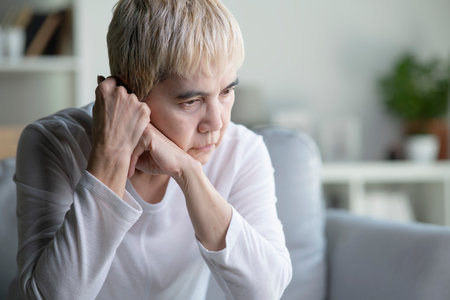 An elderly Asian woman appears sad and anxious while sitting on a sofa at home.の写真素材