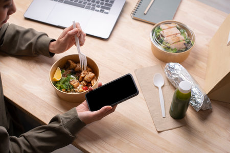 Asian woman eating healthy meal while working, Businesswoman having a lunch break at the office, hand holding phone with blank black screen for copyspaceの写真素材