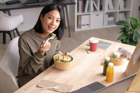 Asian woman eating healthy meal while working, Businesswoman having a lunch break at the office, Beautiful woman smiling and looking at camera while eating foodの写真素材