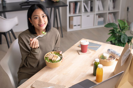 Asian woman eating healthy meal while working, Businesswoman having a lunch break at the office, Beautiful woman smiling and looking at camera while eating foodの写真素材
