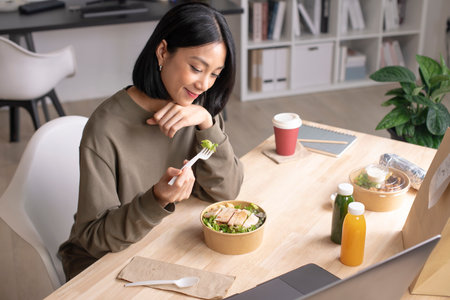 Asian woman eating healthy meal while working, Businesswoman having a lunch break at the officeの写真素材