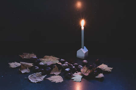 Candle on a stand that looks like a little ceramic house with autumn leaves and chestnuts around, dark backgroundの写真素材