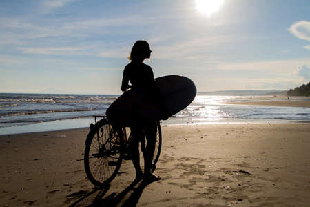 Young girl with surfboard and bicycle on the beach.の写真素材