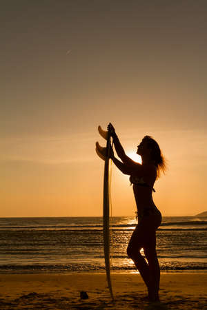 Rear view of beautiful sexy young woman surfer girl in bikini with white surfboard on a beach at sunset or sunriseの写真素材