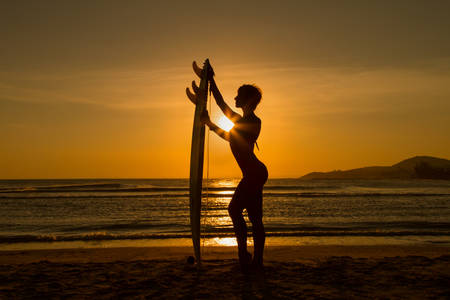 Rear view of beautiful sexy young woman surfer girl in bikini with white surfboard on a beach at sunset or sunriseの写真素材