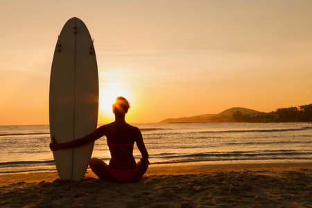Rear view of beautiful sexy young woman surfer girl in bikini with white surfboard on a beach at sunset or sunriseの写真素材