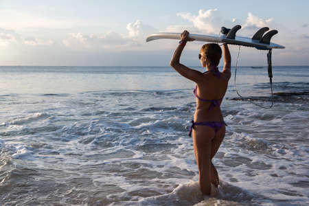 woman surfer with surfboard on tropical beach at sunset