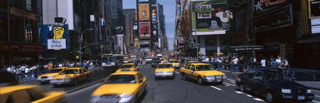 This is Times Square during the day. There are yellow taxis and oncoming traffic amidst the...