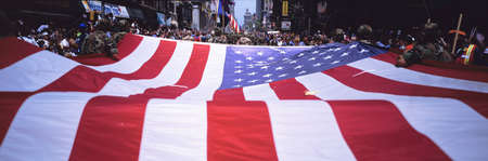 This shows a Ticker Tape Parade celebrating the Desert Storm Victory Parade in the Canyon of Heroes. The setting are buildings in New York City. There is a very large American flag stretched out lengthwise in the foreground with crowds of people surround のeditorial素材
