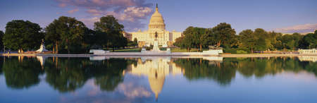 This is the U.S. Capitol showing the West side view at sunset. The Capitol is in front of the reflecting pool, showing a reflection of itself in the pool. The Capitol is surrounded by green trees and a blue sky with a public statue in front of it.のeditorial素材