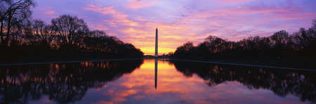 This is the Washington Monument at sunrise. The sky is a pink and purple with the monument reflected in its reflecting pool . It is surrounded by spring foliage.のeditorial素材