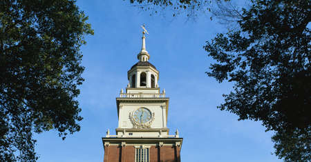 This is the top of historic Independence Hall, the site of the signing of the Declaration of Independence and the U.S. Constitution. A small dome and clock are seen toward the top. It is surrounded by green trees and blue sky.のeditorial素材