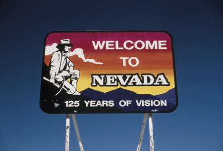 This is a road sign that says, Welcome to Nevada, 125 years of Vision. It is located on Interstate 15. It shows a miner with a pick in his hand, squatting next to the mountains against a red, orange and blue background. There is also a cloud in the sky.のeditorial素材