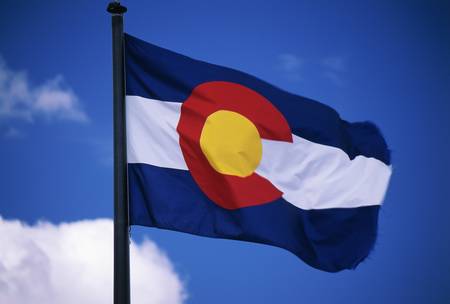 This is the Colorado State Flag, waving in the wind situated on a flag pole. It is set against a blue sky. At the center of the flag is a large capital C.の写真素材