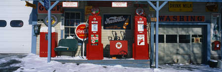 This is a vintage gas station with old style pumps that are red. It is a piece of Americana. It shows winter in New England. There is an old-fashioned Coke sign with other vintage signs. There is snow on the ground with a small bench and a garage door at のeditorial素材