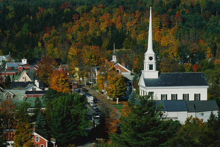 This is Scenic Route 100 in the autumn. There is a large white New England style church with a tall steeple next to smaller buildings of the town. の写真素材