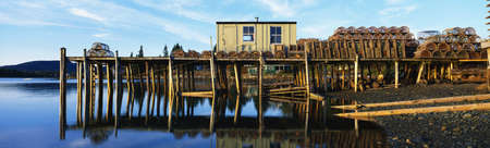 This is a pier with a small square wooden shack. There are lobster traps stacked up on the pier. The shore is in the foreground next to the water. This is also known as a Lobster Village.の写真素材