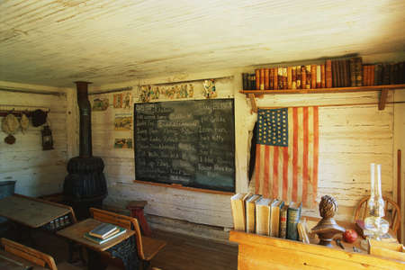 This is the interior of a one room school house. It was the first school in Montana from 1868. There is a black chalkboard and American flag hanging on the wall with a black wood furnace stove in the corner. There are a couple of old fashioned school deskのeditorial素材