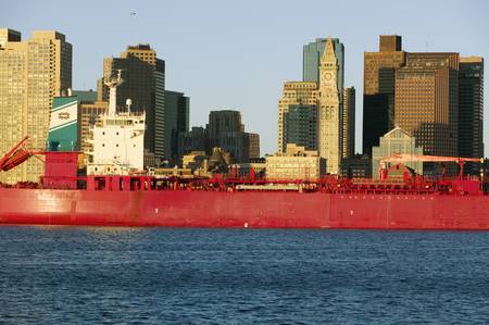 Bright red cargo ship travels in front of Boston Harbor and the Boston skyline at sunrise as seen from South Boston, Massachusetts, New Englandのeditorial素材