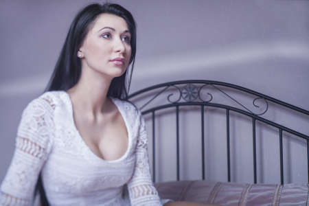 portrait of a beautiful brunette in a white dress, sitting on the bedの写真素材