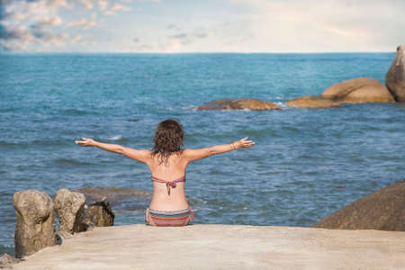 Portrait of young woman on the beach near the sea sitting with hands up. Photo from behindの写真素材