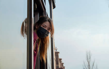 Woman with a mask on her face looks out the window at the streetの写真素材