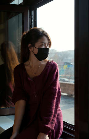 Woman with a mask on her face looks out the window at the streetの写真素材