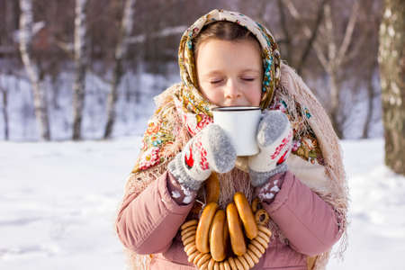 Cute girl in a traditional Russian headscarf with mug of hot tea and bagels on winter background. Closeup portrait of a child in folk clothes. Maslenitsa festival.の写真素材