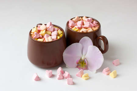 Brown cups of hot cocoa with marshmallows in heart shape on white background. Romantic background with hot chocolate and orchid flower. Love concept for Valentine's Day or wedding.の写真素材
