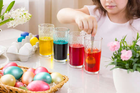 Cute little girl painting Easter eggs in glasses with liquid dye at home. Happy child preparing for Easter day. Focus picked.の写真素材