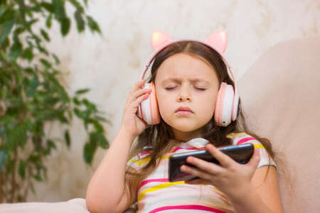 Cute little girl in headphones is using a smartphone and listening to music at home. selective focus. close-up portrait. Kid using technology.の写真素材