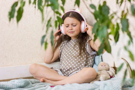 Cute little girl with wireless headphones and smartphone sitting on the sofa at home.の写真素材
