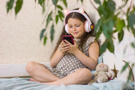 Cute little girl with wireless headphones and smartphone sitting on the sofa at home.の写真素材