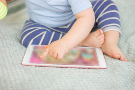 Cute toddler baby boy sitting on the bed at home and playing with digital tablet. Closeup kid hand touching screen device. child using modern technology. early development. Focus picked.の写真素材