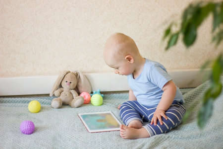 Cute toddler baby boy sitting on the bed at home, using digital tablet and watching cartoons. Child...
