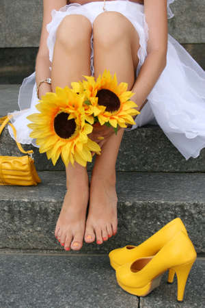 Yellow shoes and handbag behind at the feet of the bride, who is sitting and holding hands in sunflowersの写真素材