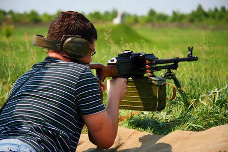 A man in a dark shirt and headphones lies with a machine gun and shoots