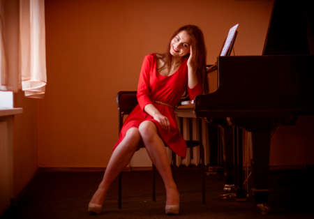 Happy woman in red dress sitting near grand piano, smiling and looking at cameraの写真素材