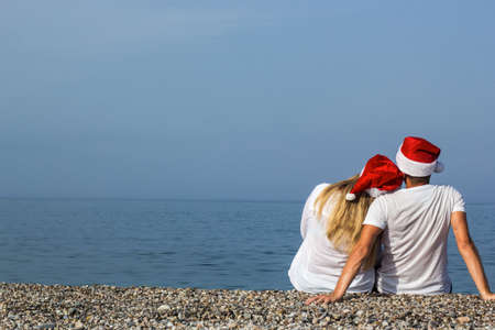 Back view of romantic couple in red Santa hats sitting on beachの写真素材
