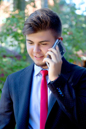 Young college student standing at park and calling on his mobile phone. Teenager dressing in suit,...
