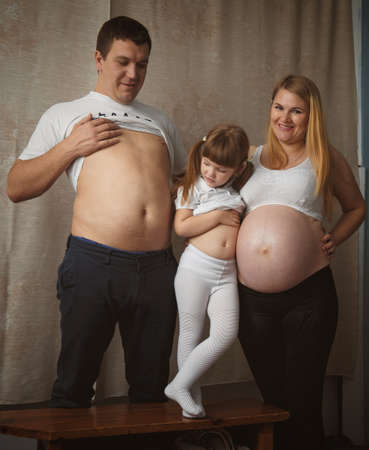 Young man, little girl and pregnant woman showing their big bellies at homeの写真素材