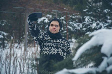 Young man in scandinavian coat and hat taking selfie picture with smartphone at winter parkの写真素材