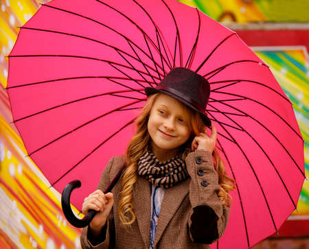 Adorable, elegant school aged kid girl wearing jacket and hat holding pink umbrella walking in the park autumn dayの写真素材