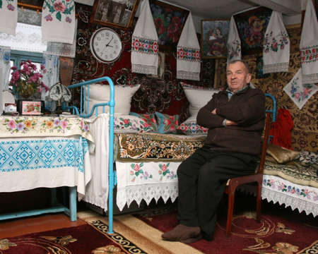 NOVA VOLIA, UKRAINE - 14 November 2008: Unknown senior man sits near bed with embroidered pillows in rural parlorのeditorial素材